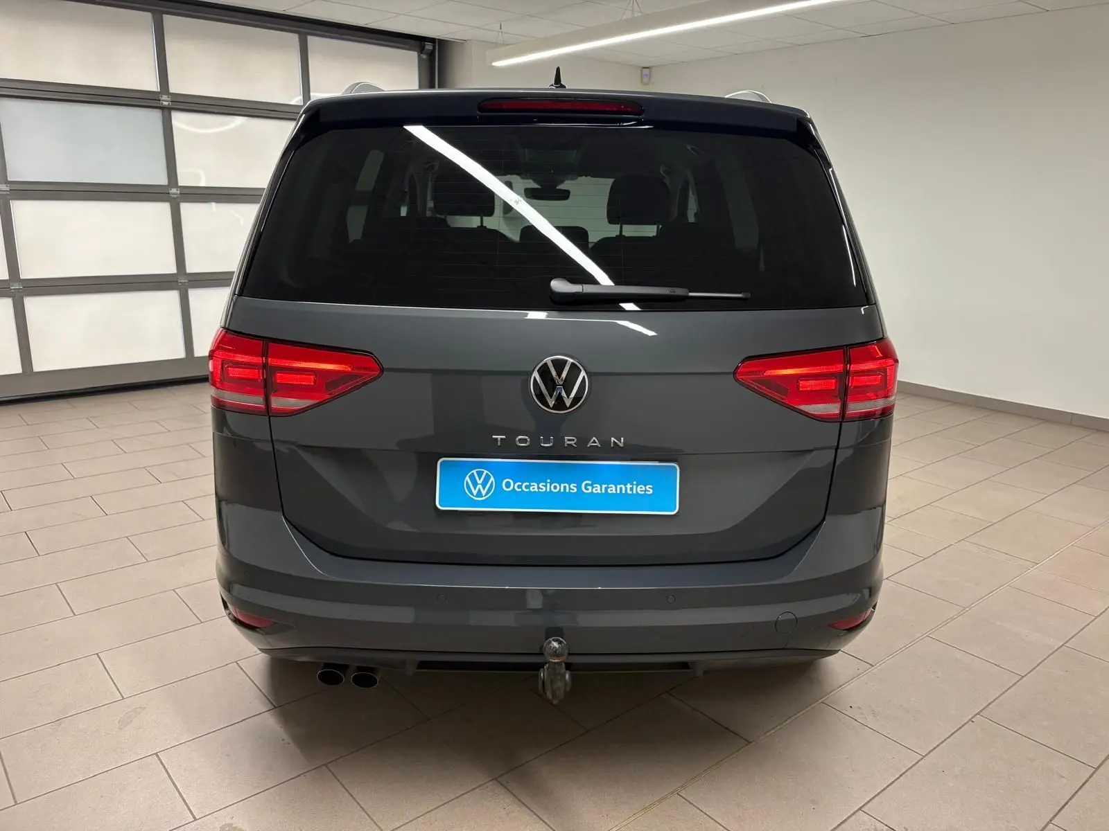 Vue arrière d'un Volkswagen Touran gris dauphin avec attelage escamotable et feux rouges allumés en intérieur.