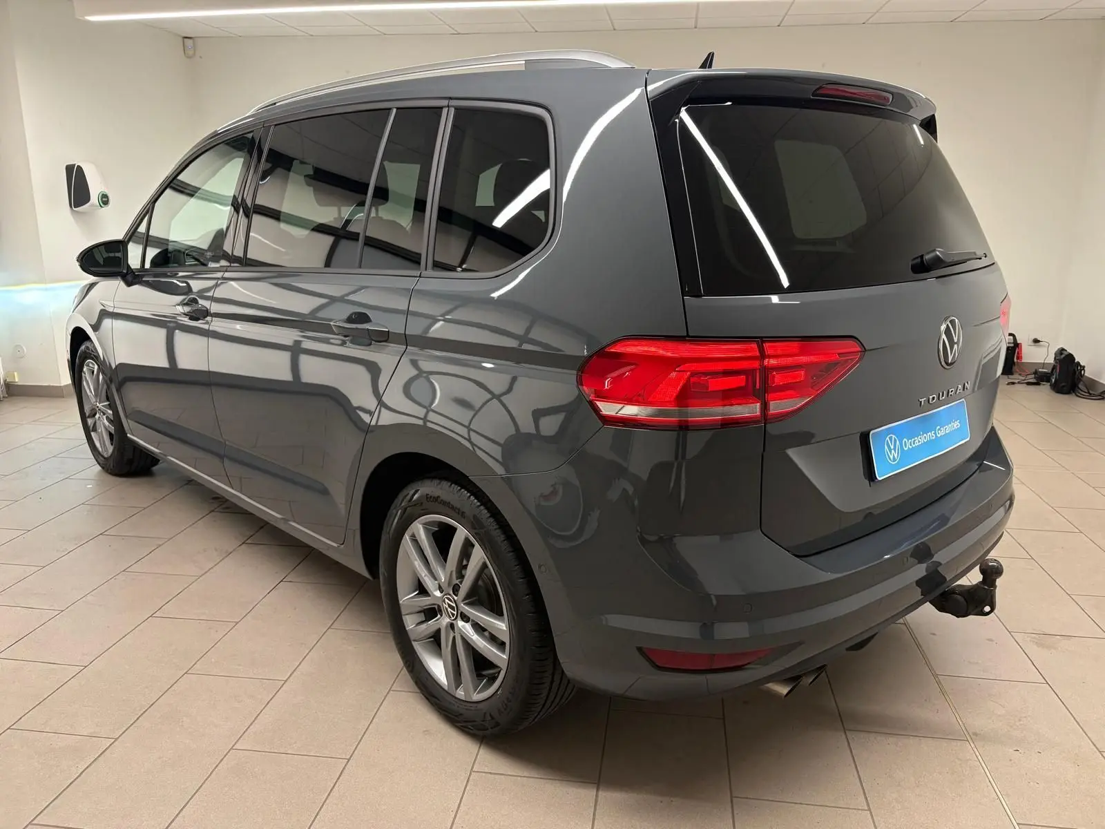 Vue 3/4 arrière droite d’un Volkswagen Touran gris dauphin avec attelage escamotable et vitres teintées.