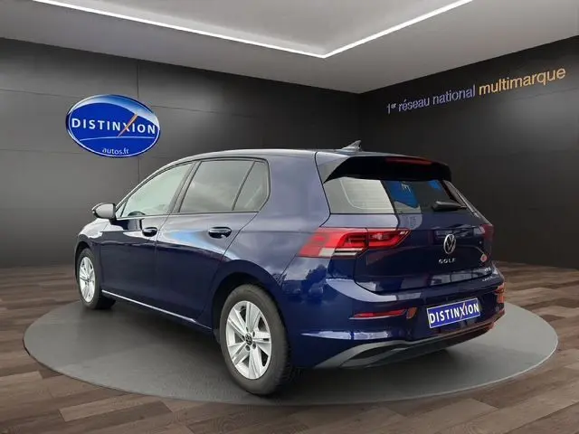 Volkswagen Golf VIII bleu en 3/4 arrière droit, avec hayon fermé et feux arrière allumés en showroom.