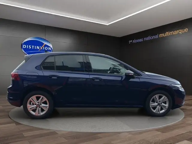 Profil droit d'une Volkswagen Golf VIII bleu foncé 5 portes exposée en showroom avec jantes alliage et vitres teintées.