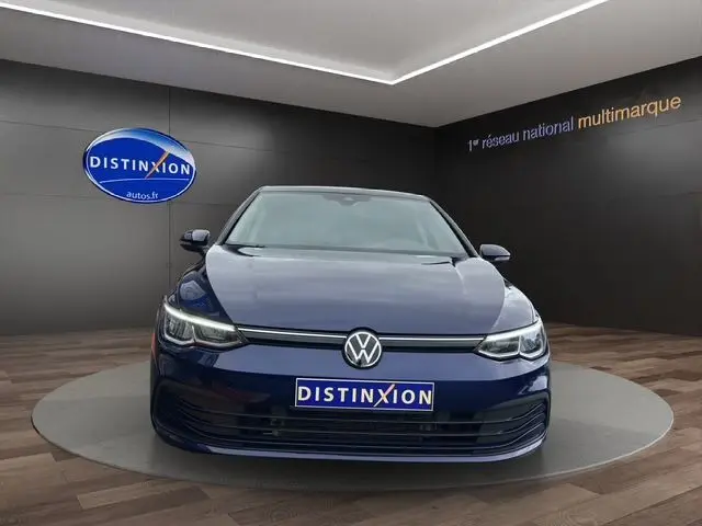 Vue frontale d'une Volkswagen Golf VIII bleu foncé avec phares LED et logo VW bien visible en showroom.