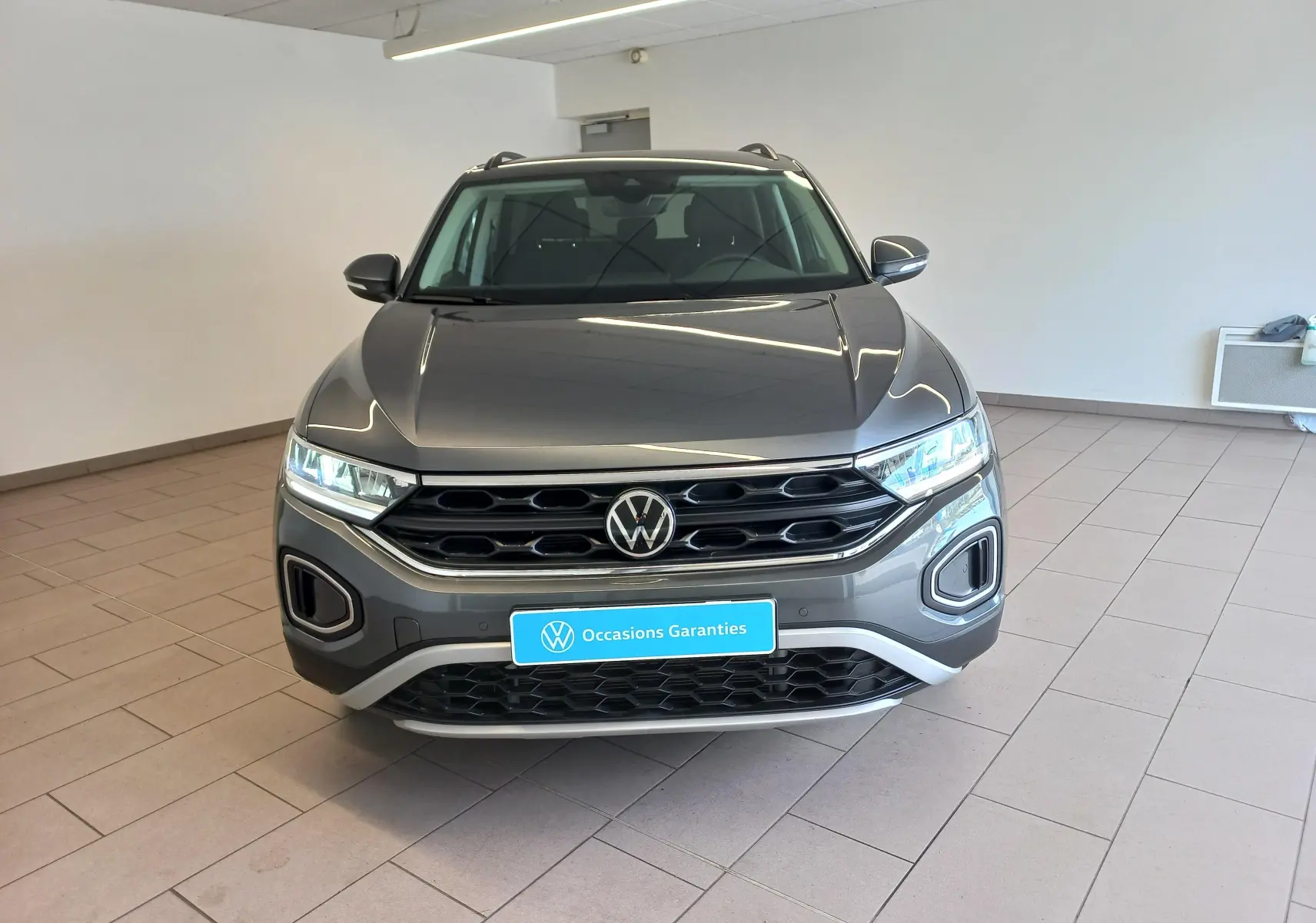 Vue avant d’un Volkswagen T-Roc 1.5 TSI 150 DSG7 VW Edition gris Indium avec calandre noire et feux LED allumés.