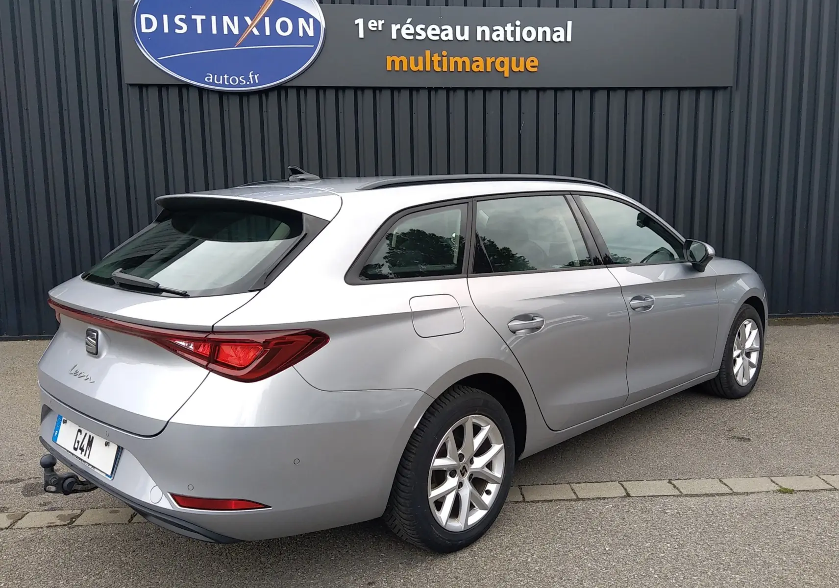 Vue 3/4 arrière droite d’une SEAT LEON ST gris métallisé avec attelage, garée devant un mur gris.