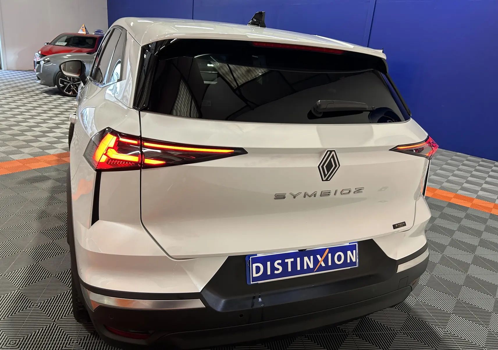 Vue arrière droite d'une Renault SYMBIOZ blanc nacré avec feux LED allumés et hayon électrique visible en intérieur showroom.