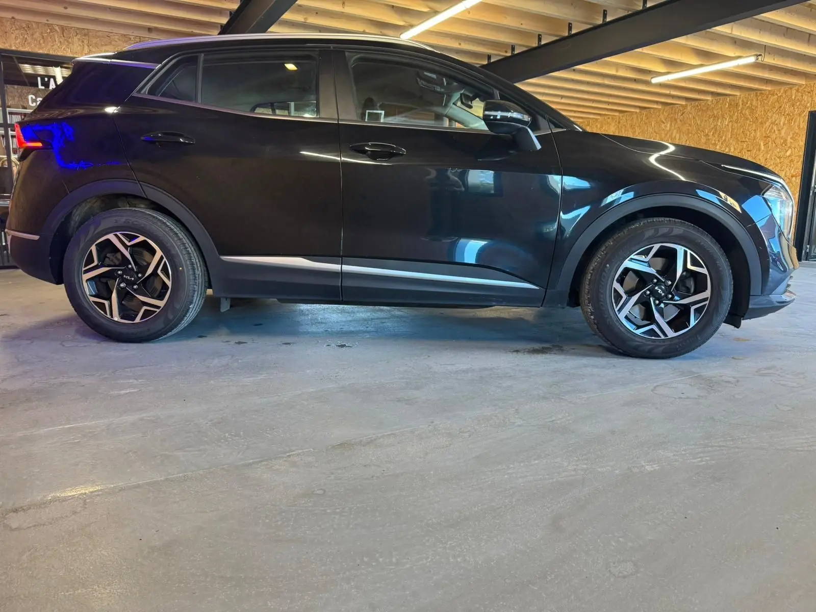 Profil côté gauche d’un KIA Sportage noir 2022 avec jantes alliage et détails chromés sous un plafond en bois.