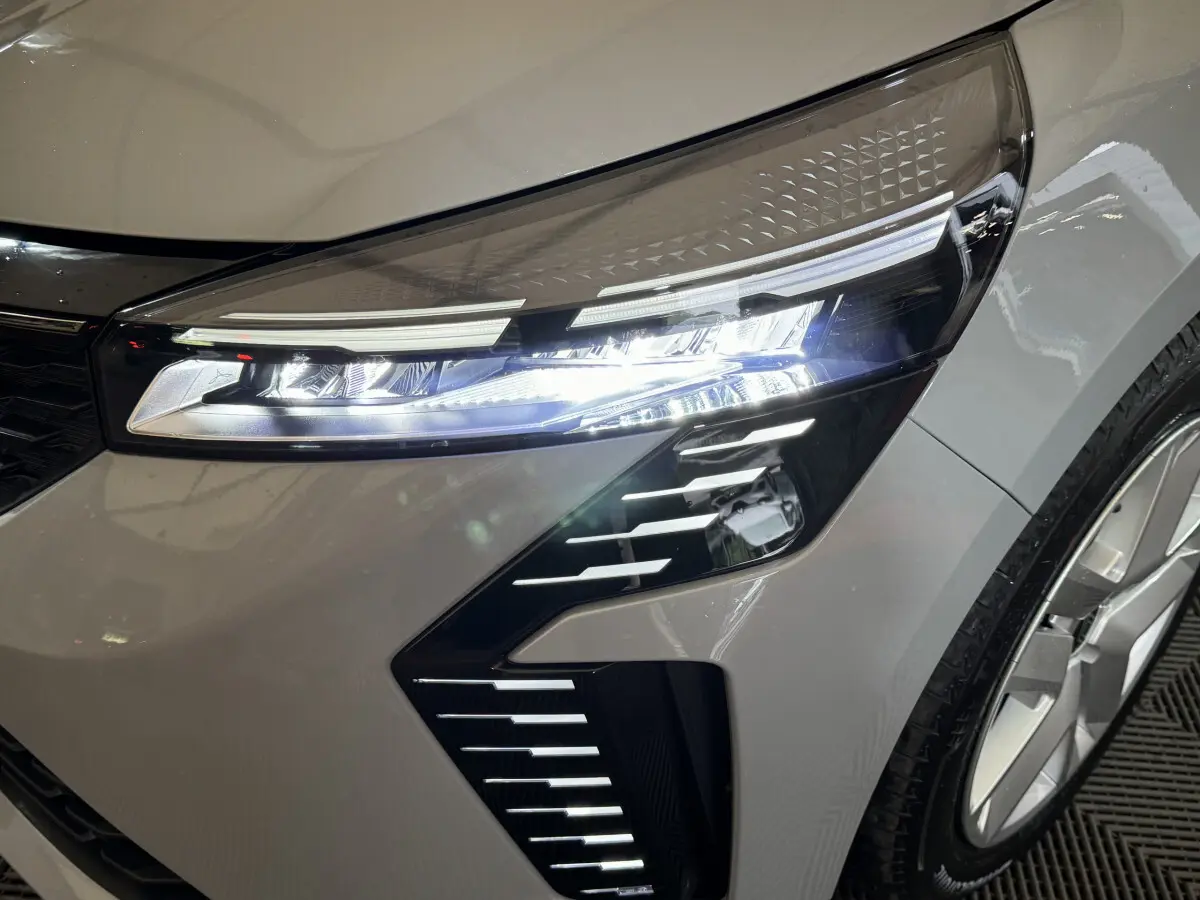 Gros plan sur le phare avant droit allumé d'une Renault Clio gris clair, avec jante et partie de la calandre visibles.