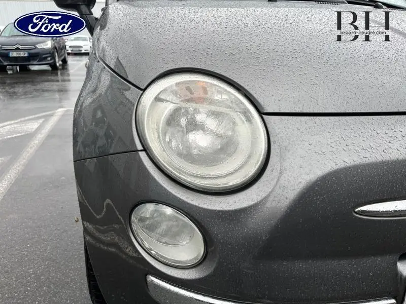 Gros plan sur le phare avant gauche d'une FIAT 500 marron sous la pluie, avec gouttes d'eau visibles sur la carrosserie.
