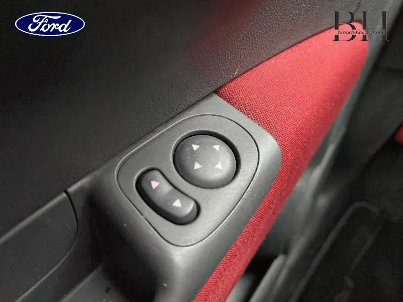 Gros plan sur les commandes électriques des rétroviseurs côté conducteur, sur panneau intérieur rouge d'une Fiat 500 marron.