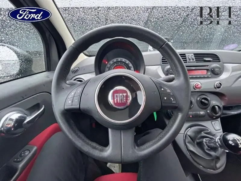 Vue intérieure centrée sur le volant noir avec logo FIAT, tableau de bord gris et levier de vitesse d'une Fiat 500 marron.