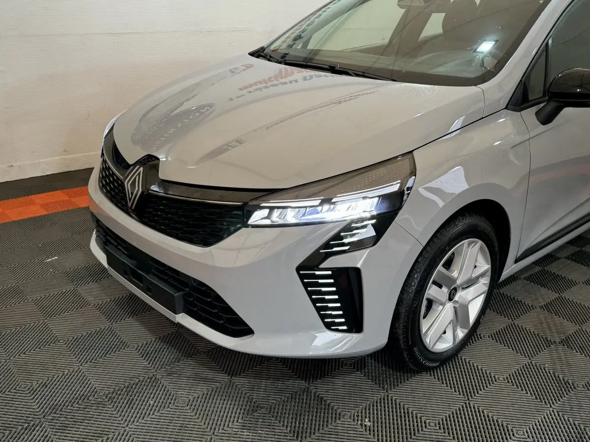 Vue 3/4 avant droit d'une Renault Clio gris clair avec phares LED allumés et calandre noire brillante.