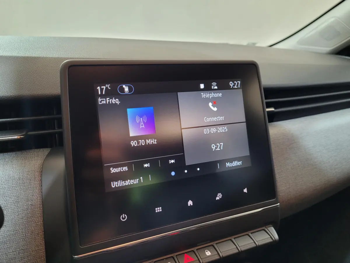 Écran tactile central de la Renault Clio gris foncé, affichant la radio et la connexion téléphone, vue intérieure frontale.