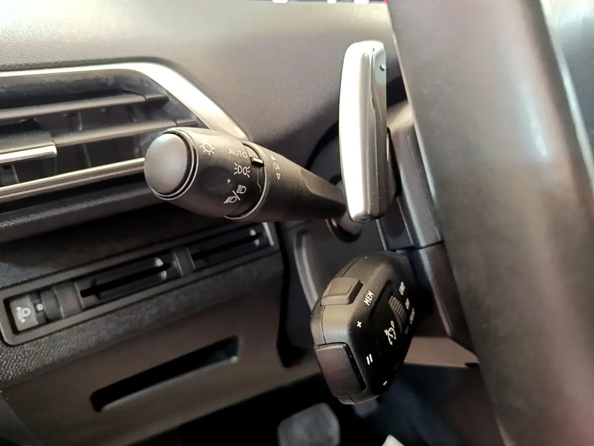 Gros plan sur la commande des phares et le volant cuir noir à gauche du tableau de bord du Peugeot 5008 gris clair.