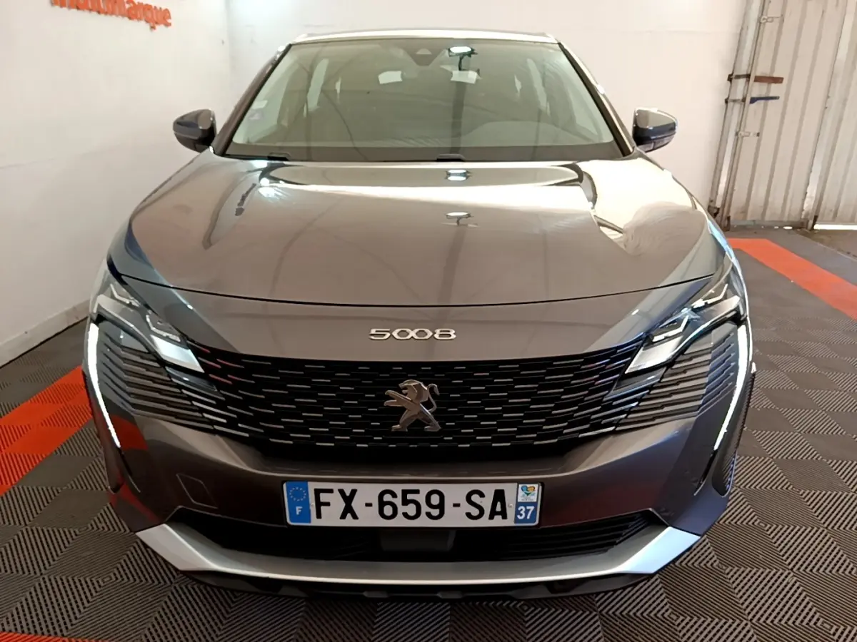 Peugeot 5008 gris clair vue de face, avec calandre noire et phares LED allumés dans un garage.