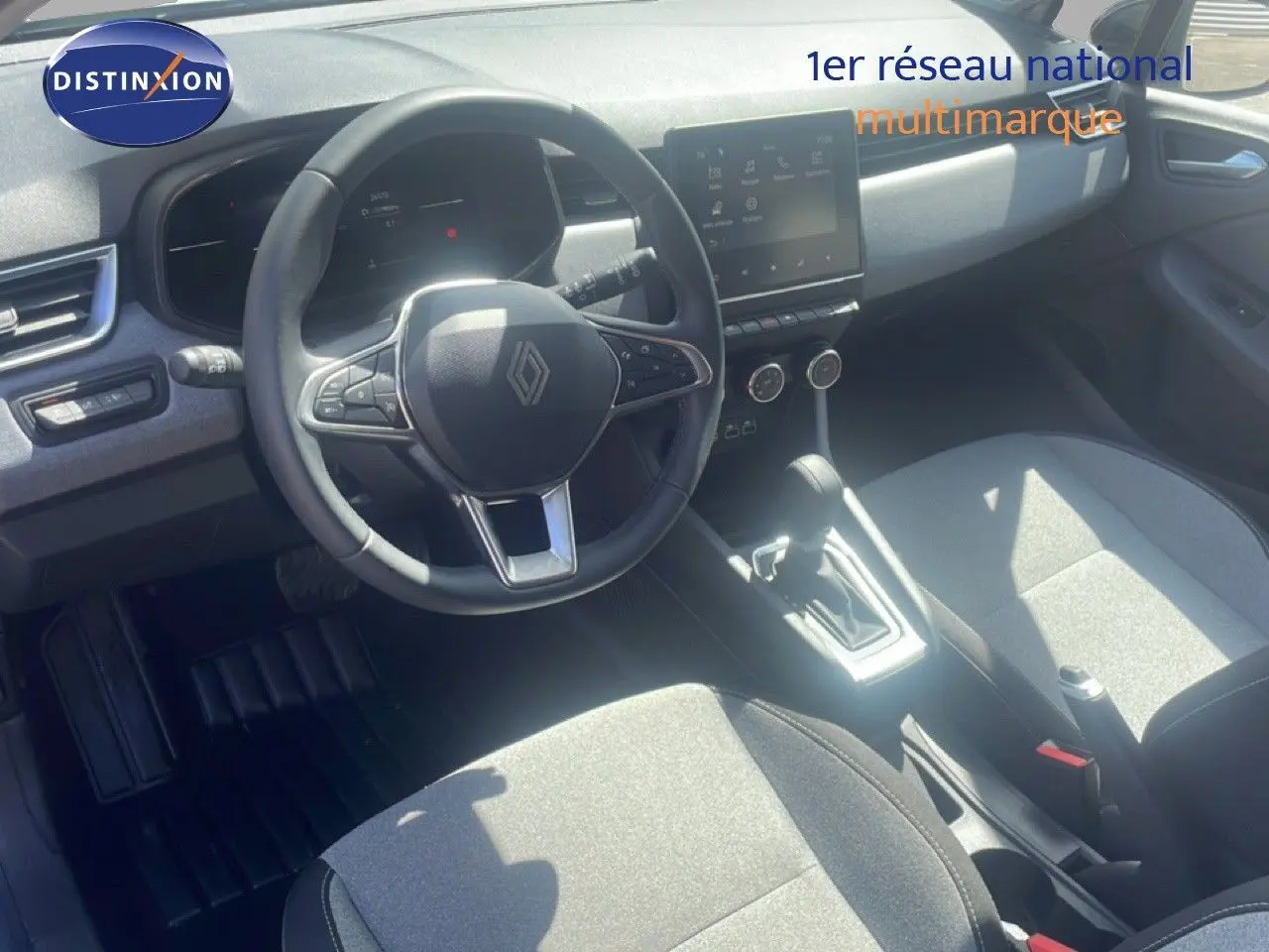 Vue intérieure de la Renault Clio 2025 montrant le volant, la console centrale avec écran tactile et les sièges tissu gris.