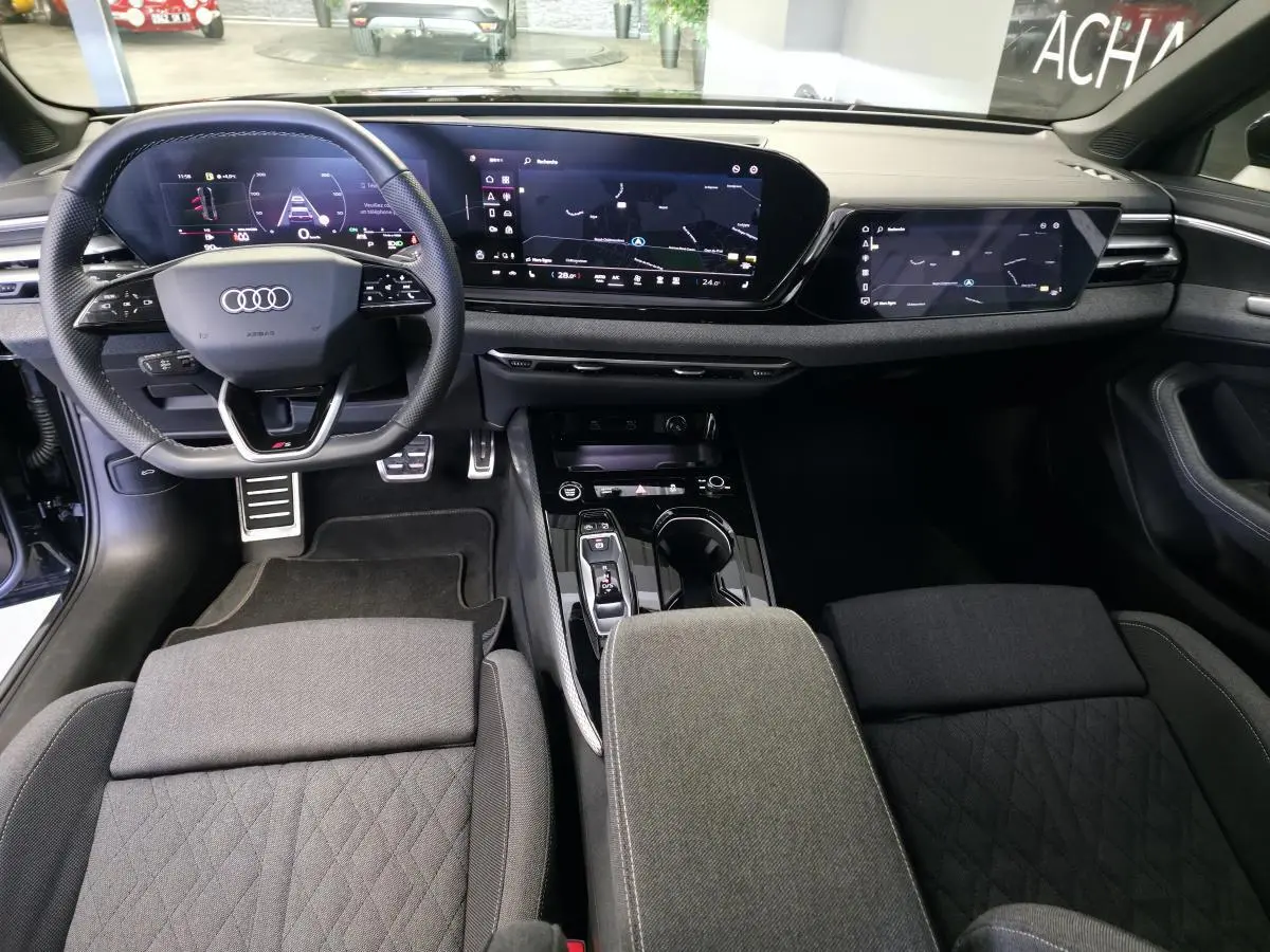 Vue intérieure avant de l'Audi A5 2025 S line, tableau de bord noir avec double écran numérique et volant sport cuir.