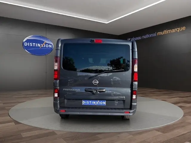 Vue arrière d'un Nissan Primastar gris foncé, avec vitres teintées et feux arrière verticaux visibles en intérieur showroom.