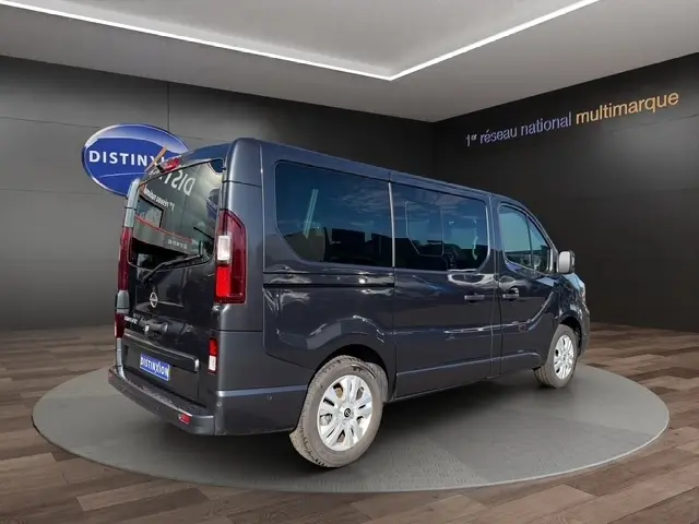 Vue 3/4 arrière droite d’un Nissan Primastar gris foncé avec vitres teintées et jantes argentées, en showroom.