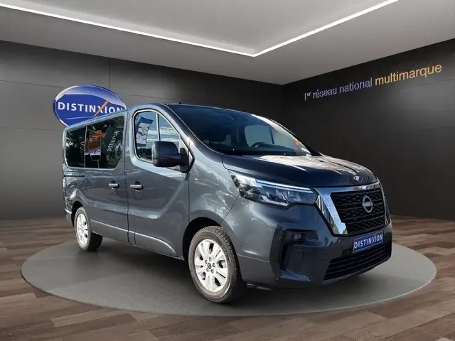 Vue 3/4 avant d’un Nissan Primastar gris foncé en showroom, avec calandre noire et jantes argentées visibles.