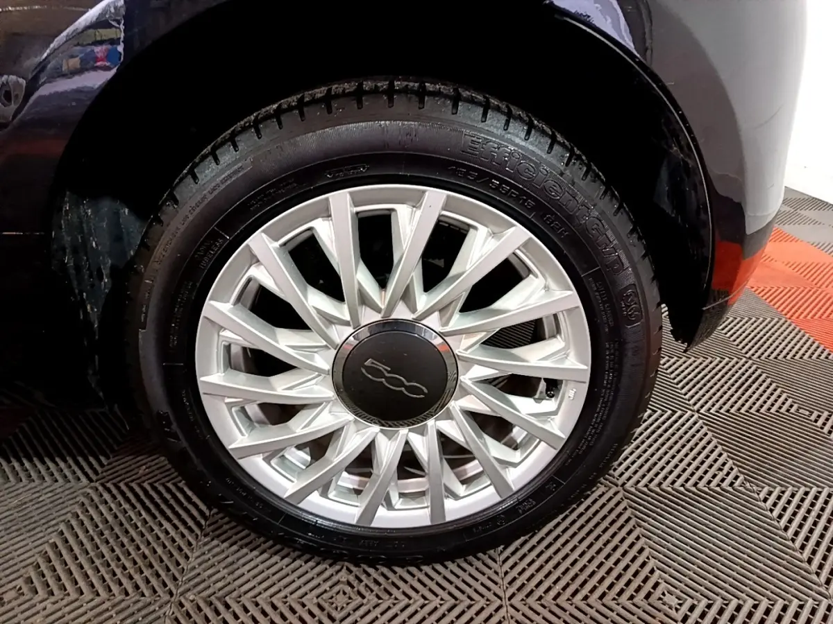 Gros plan sur la roue avant droite d'une FIAT 500 noire, mettant en valeur la jante argentée au design étoilé.