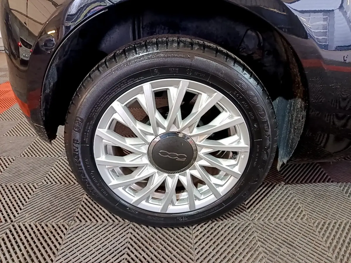 Gros plan sur la roue avant droite d'une FIAT 500 noire, mettant en valeur la jante argentée au design complexe.