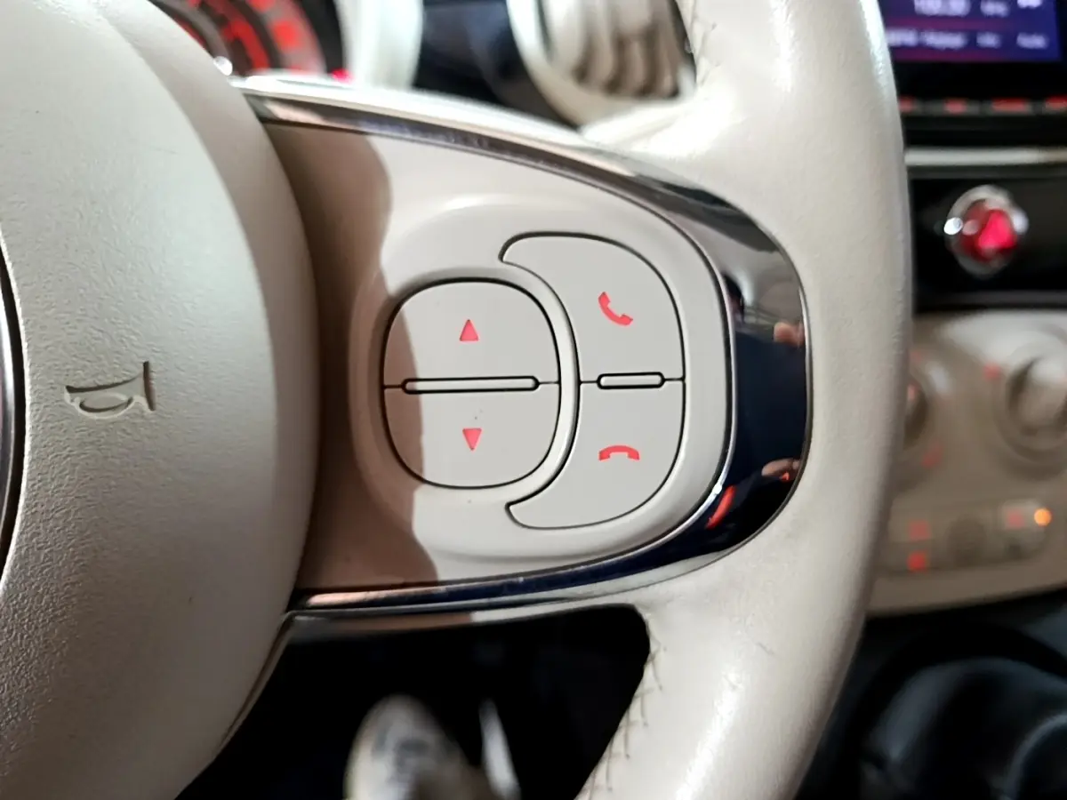 Gros plan sur les commandes blanches du volant cuir beige d'une Fiat 500 Lounge 2019, avec tableau de bord flou en arrière-plan.