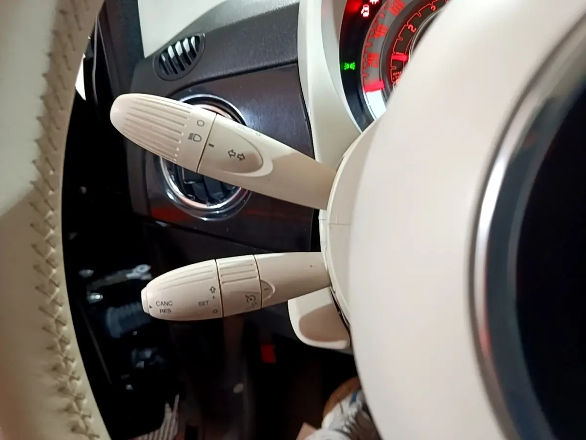 Gros plan sur les commandes de clignotants et régulateur au volant beige d’une Fiat 500 Lounge noire, tableau de bord partiellement visible.