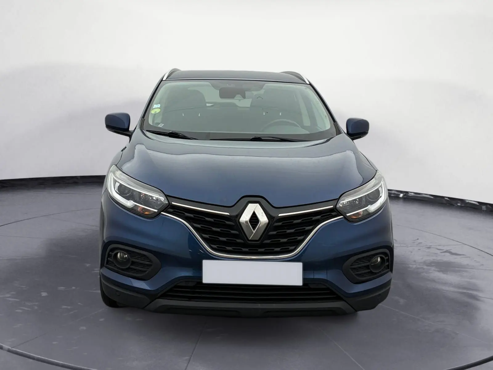 Renault Kadjar bleu cosmos en 3/4 avant droit sur un parking extérieur avec plusieurs fourgons blancs en arrière-plan.