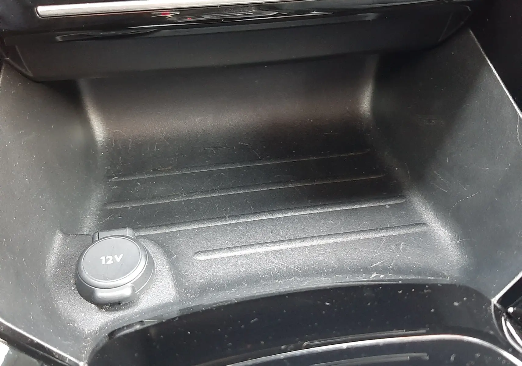 Gros plan sur la prise 12V et le rangement central noir brillant de la Peugeot 2008 Hybrid 145 Allure 2025.