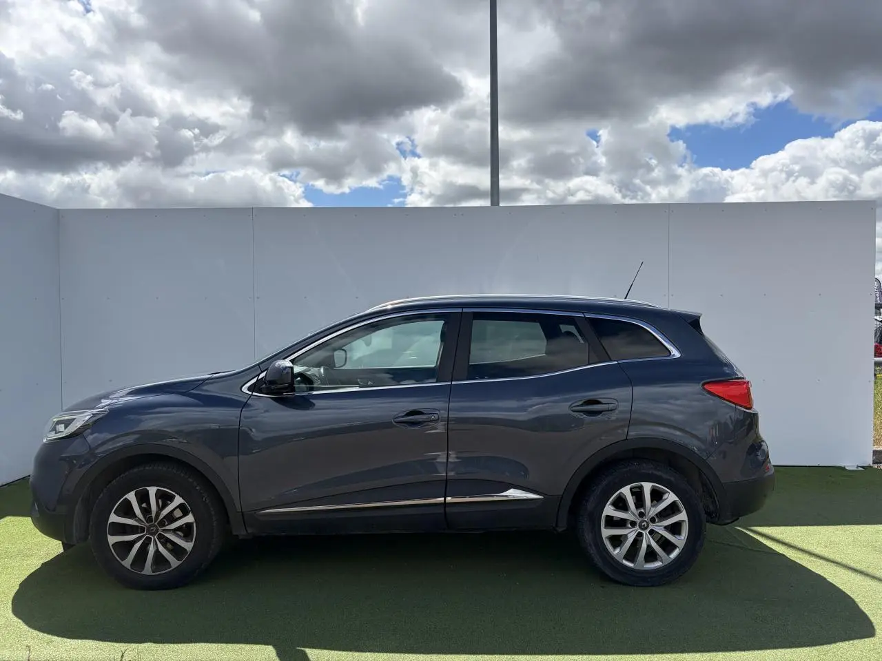 Profil côté gauche d'un Renault Kadjar 2016 gris titanium avec jantes alliage 17 pouces et barres de toit chromées.