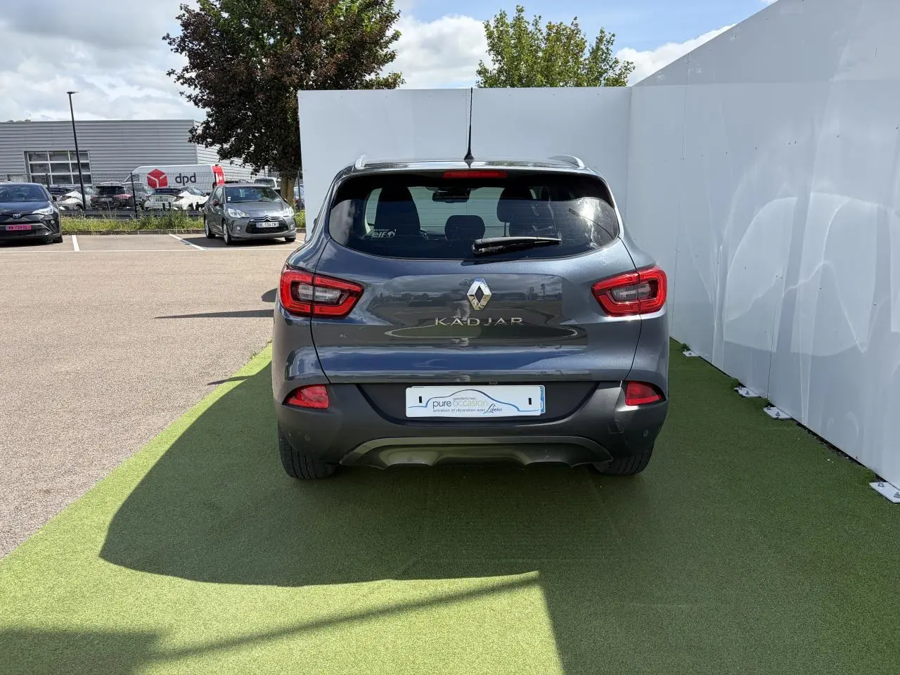 Vue arrière d'un Renault Kadjar bleu métal garé sur sol vert avec feux arrière rouges et logo chromé visible.