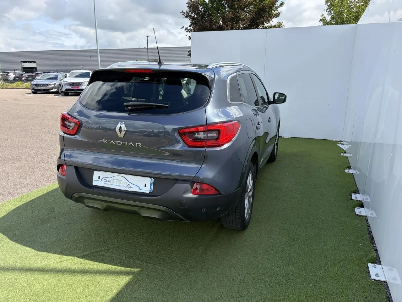 Vue 3/4 arrière droite d'un Renault Kadjar bleu métal garé sur sol vert, avec feux arrière et logo visibles.
