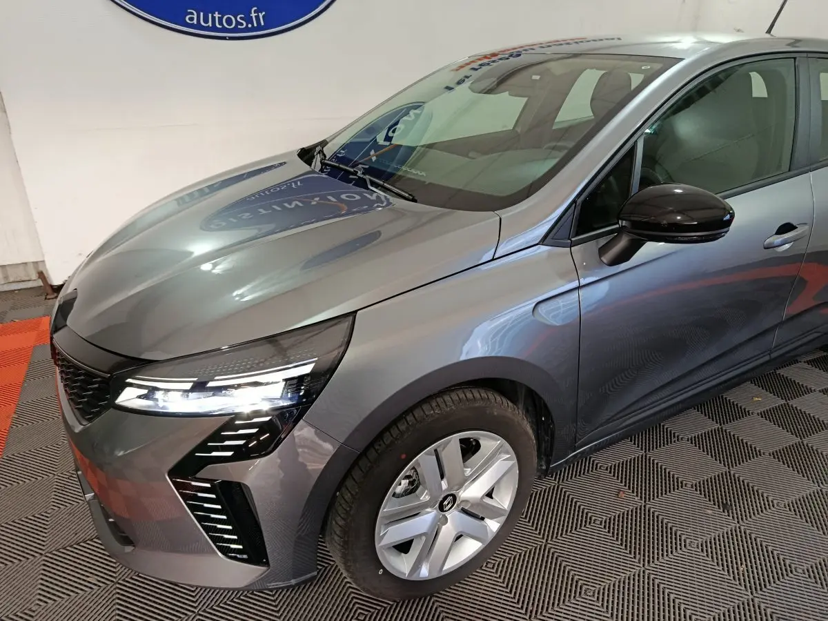 Renault Clio gris clair vue 3/4 avant droit, avec phares LED allumés et rétroviseur noir brillant.