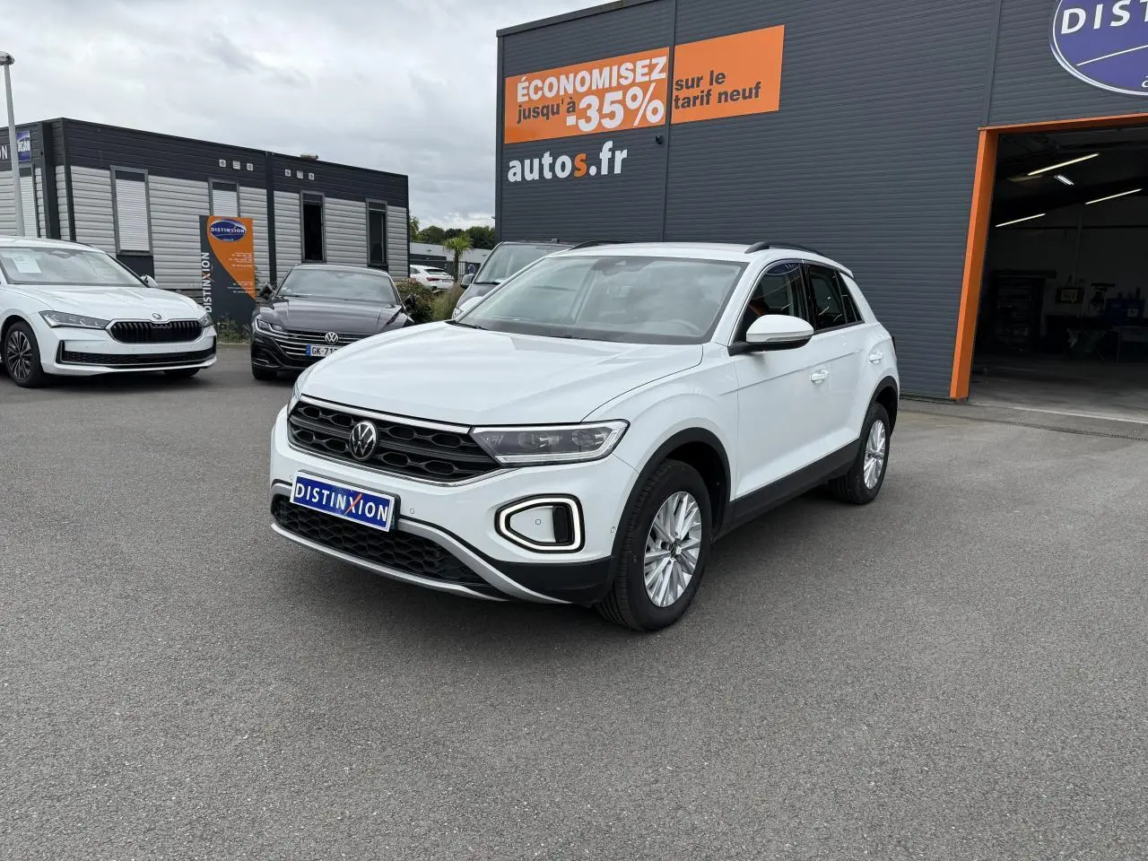 Volkswagen T-Roc blanc pur vu en 3/4 avant droit sur parking devant un garage avec panneau promotionnel orange.