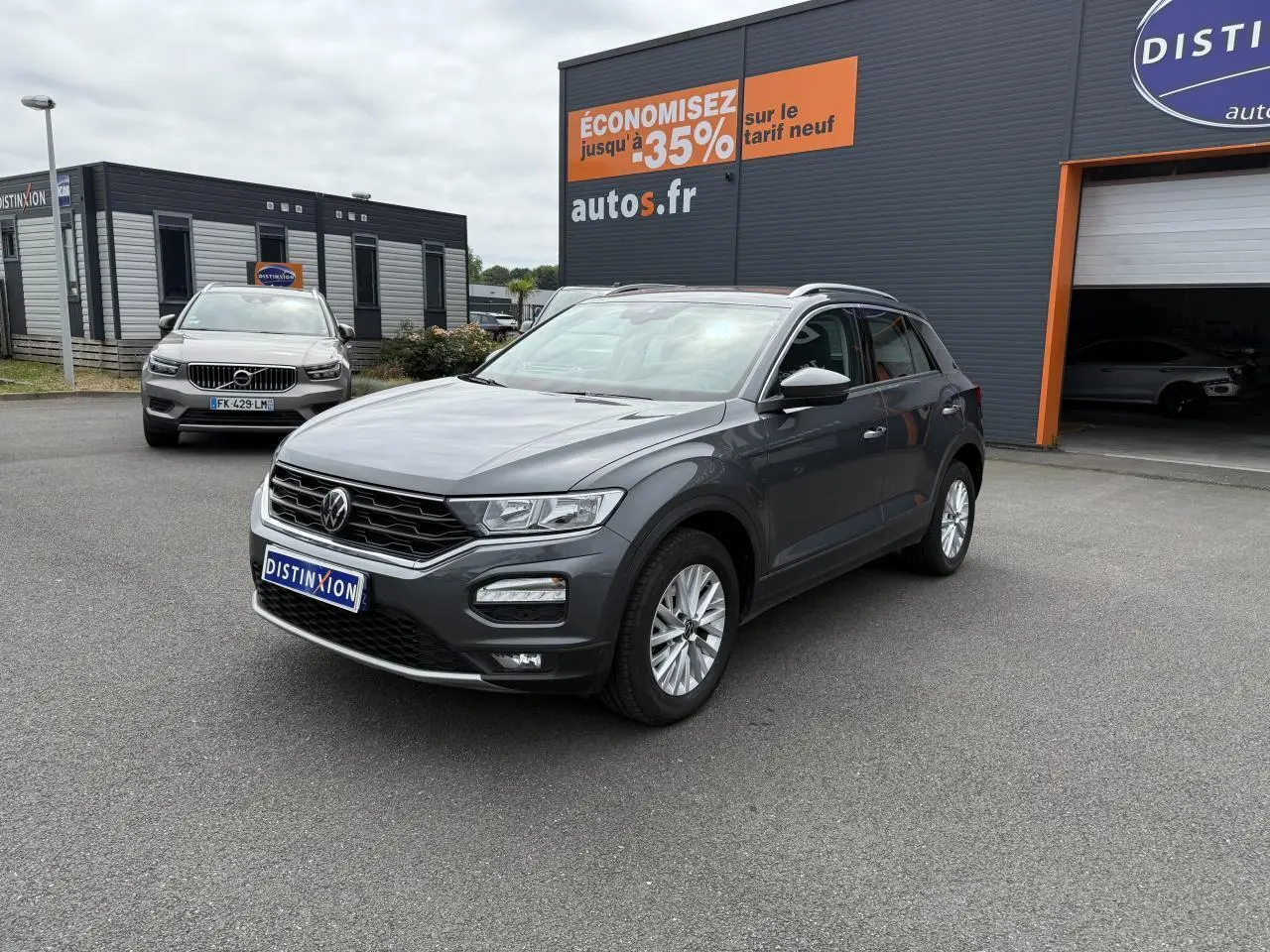 Volkswagen T-Roc gris Indium Metal en 3/4 avant droit, stationné devant un garage avec panneaux publicitaires.