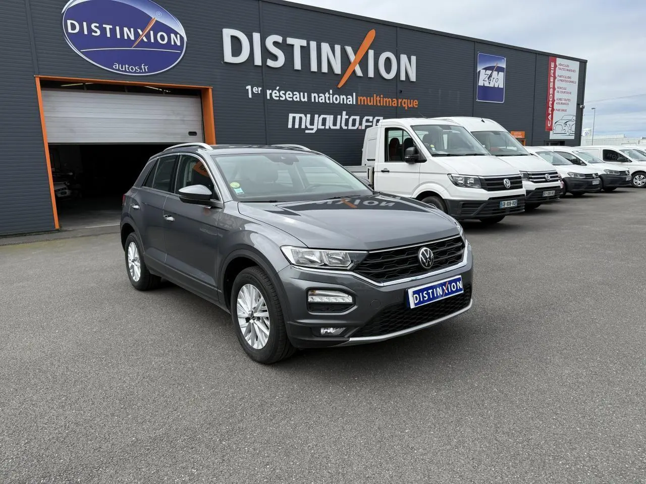 Volkswagen T-Roc gris Indium Metal en 3/4 avant droit devant un bâtiment Distinxion avec plusieurs véhicules utilitaires blancs.