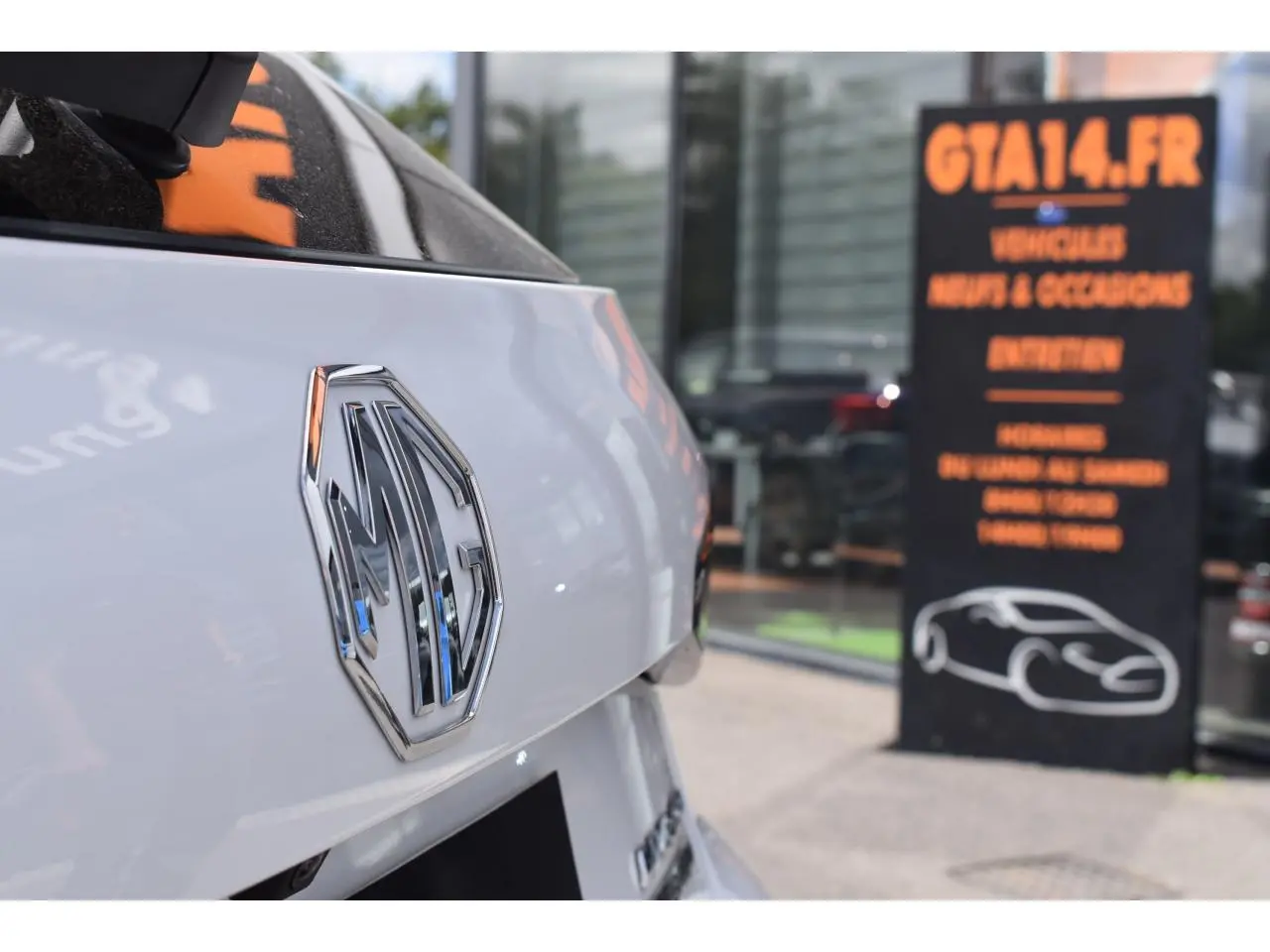 Gros plan sur l'arrière blanc brillant d'une MG3 1.5L Hybrid+ avec logo MG chromé, devant une vitrine commerciale.