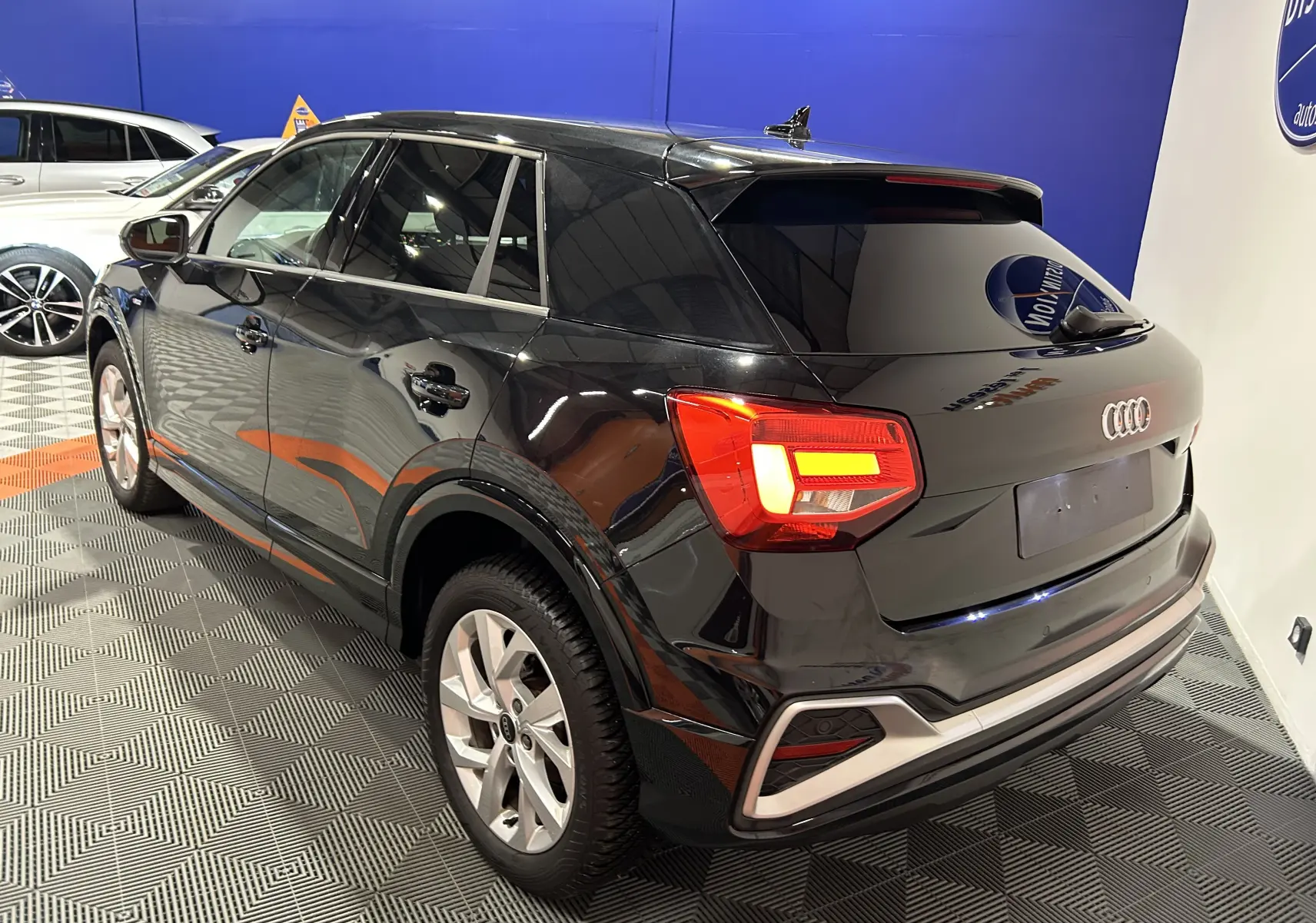 Audi Q2 noir métal vue 3/4 arrière droit, hayon électrique et feux arrière LED allumés dans un showroom.