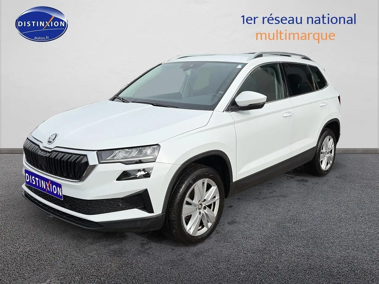Vue 3/4 avant d'un Skoda Karoq blanc glacial avec jantes alu et barres de toit noires sur fond neutre.
