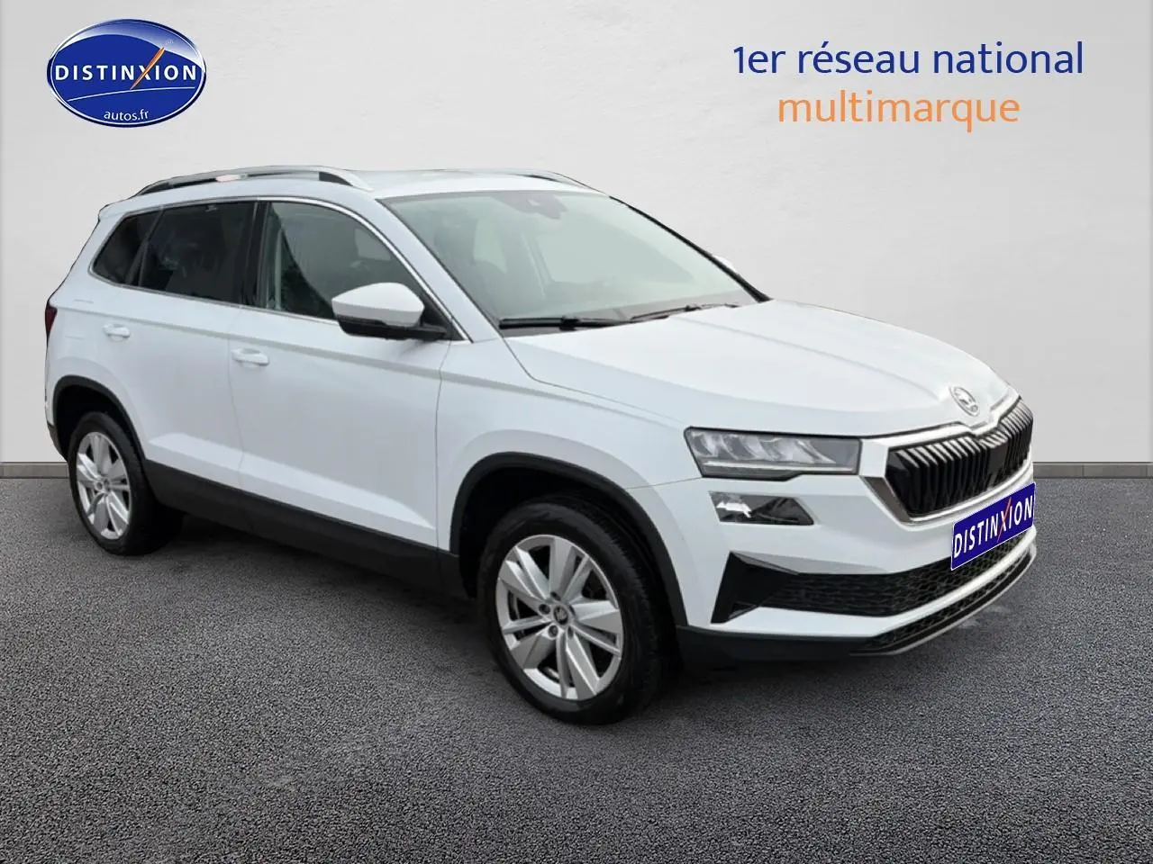 Vue 3/4 avant d'un Skoda Karoq blanc glacial avec jantes alu et barres de toit noires sur fond neutre.