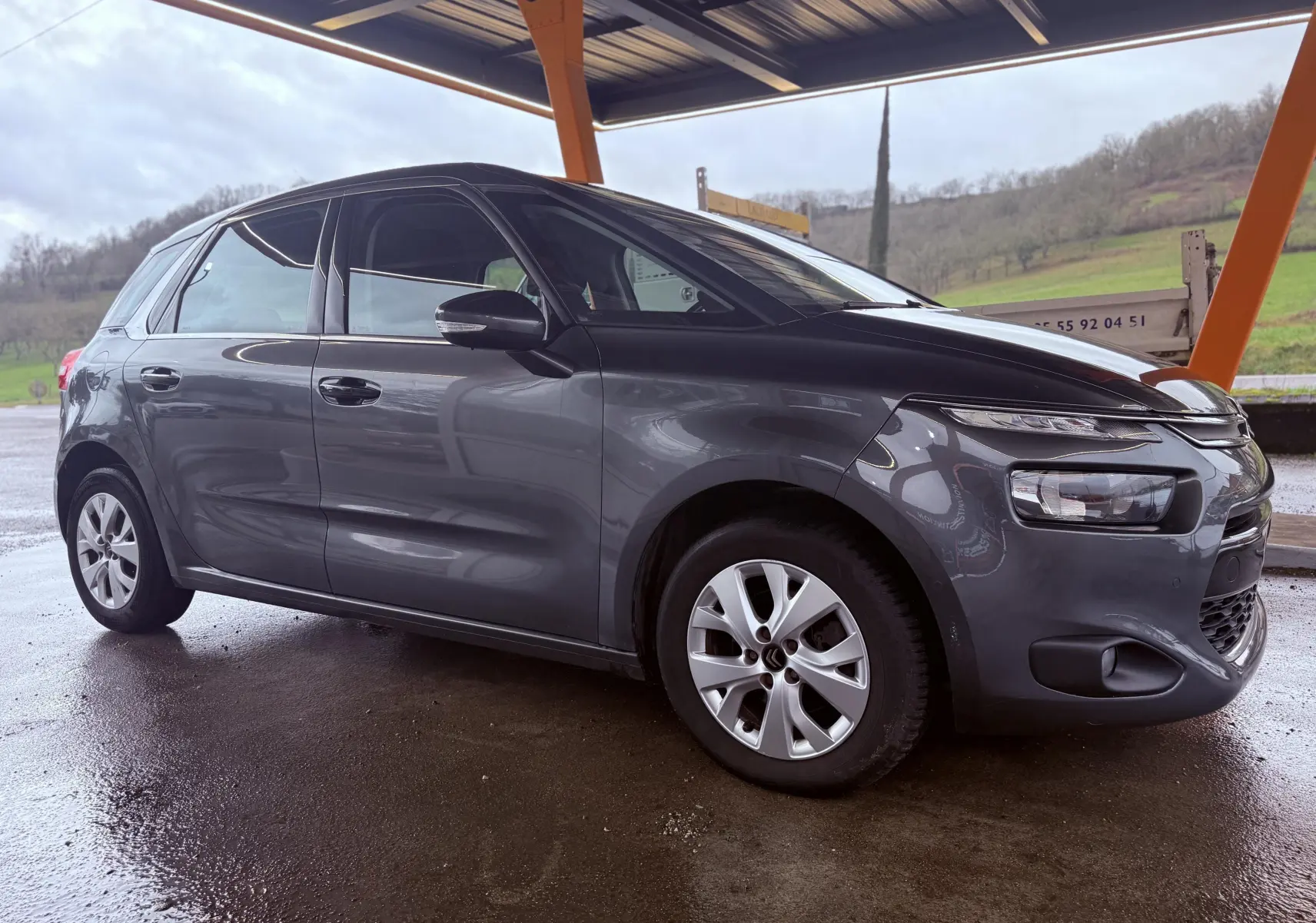 Vue 3/4 avant droit d'une Citroën C4 Picasso gris platinium métal sous abri, avec jantes alliage et phares distinctifs.