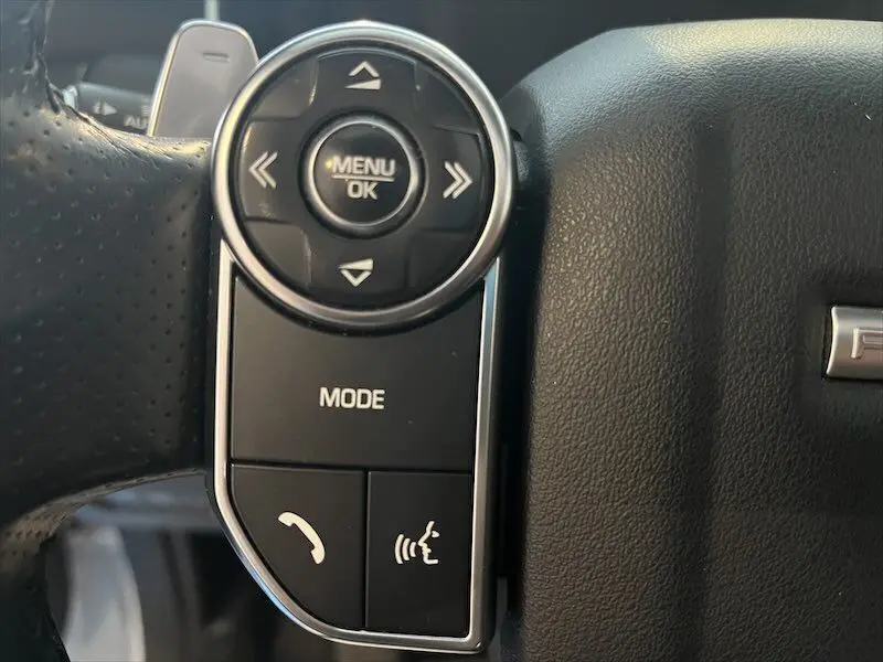 Gros plan sur les commandes au volant noir du Range Rover Sport blanc, avec boutons de menu et téléphone visibles.