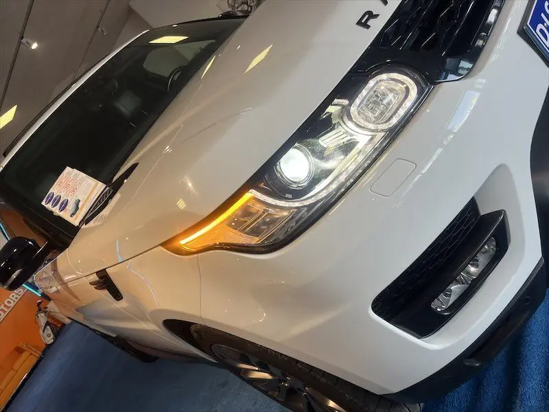 Vue 3/4 avant droit d'un Range Rover Sport blanc avec phares LED allumés dans un showroom.