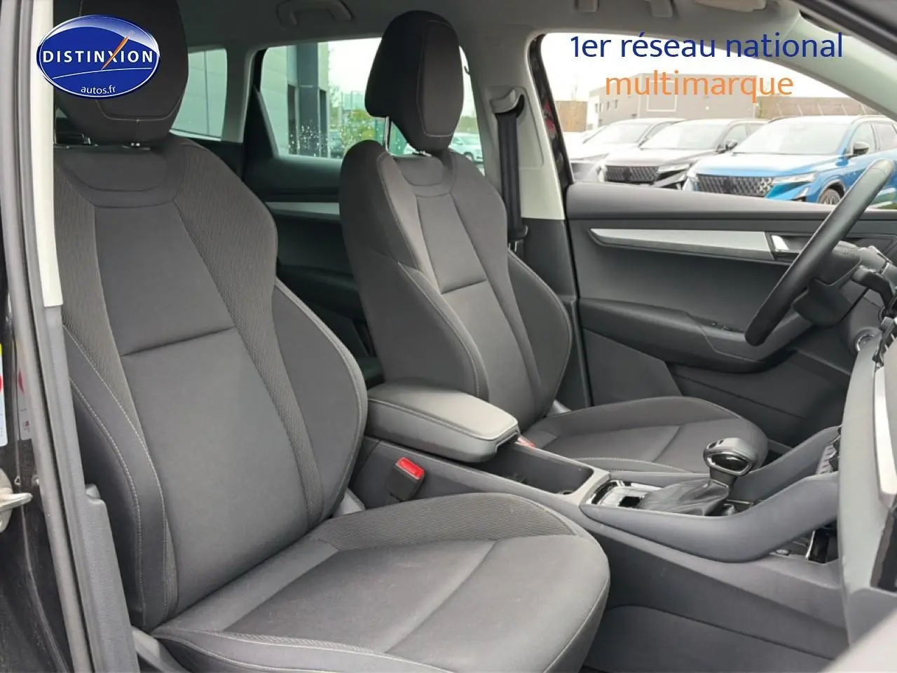Vue intérieure côté avant droit du Skoda Karoq noir 2025, montrant sièges tissu gris et accoudoir central.