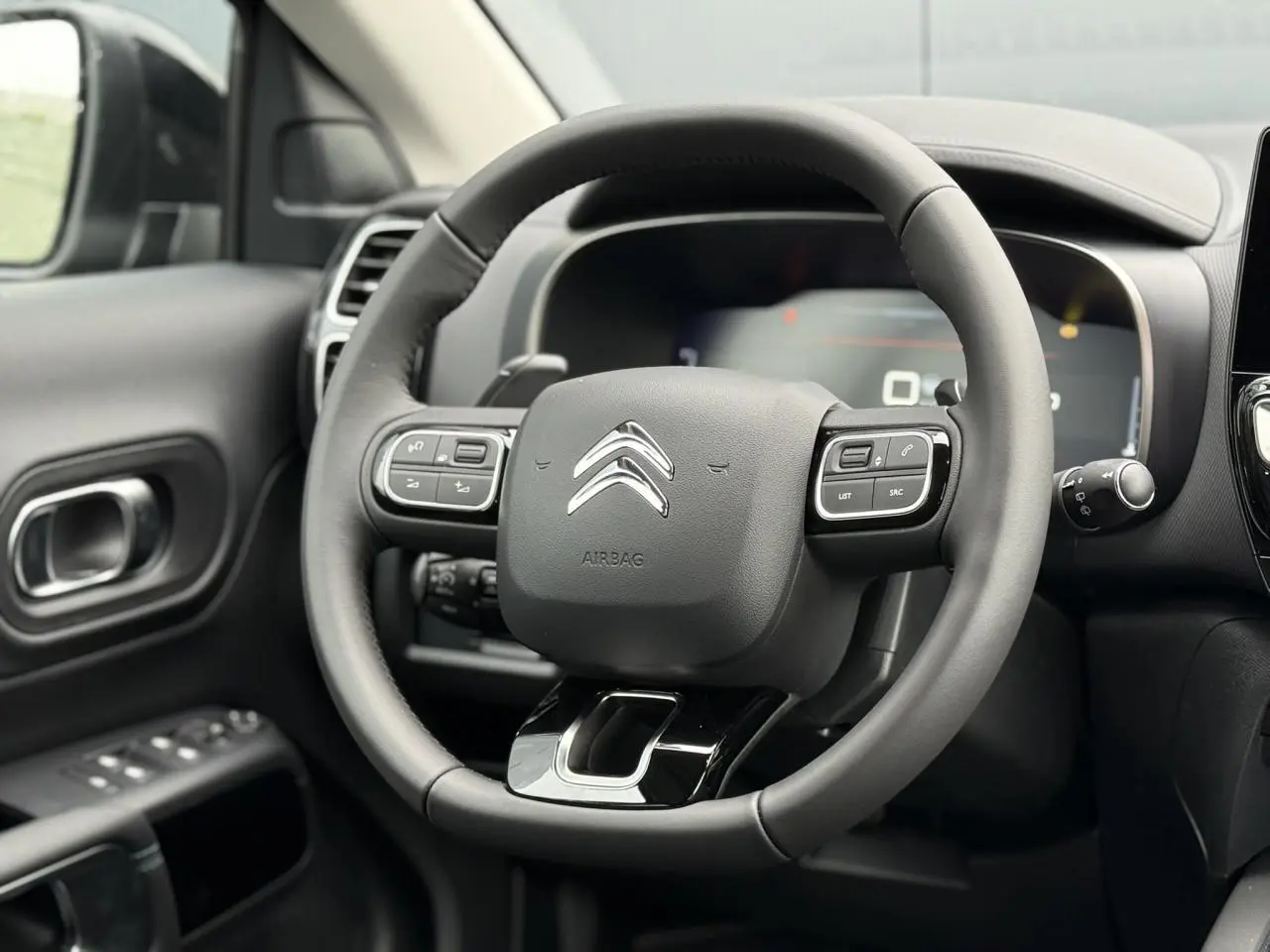 Volant noir de Citroën C5 Aircross Hybrid 2025 vu de face, avec commandes intégrées et tableau de bord numérique flou.