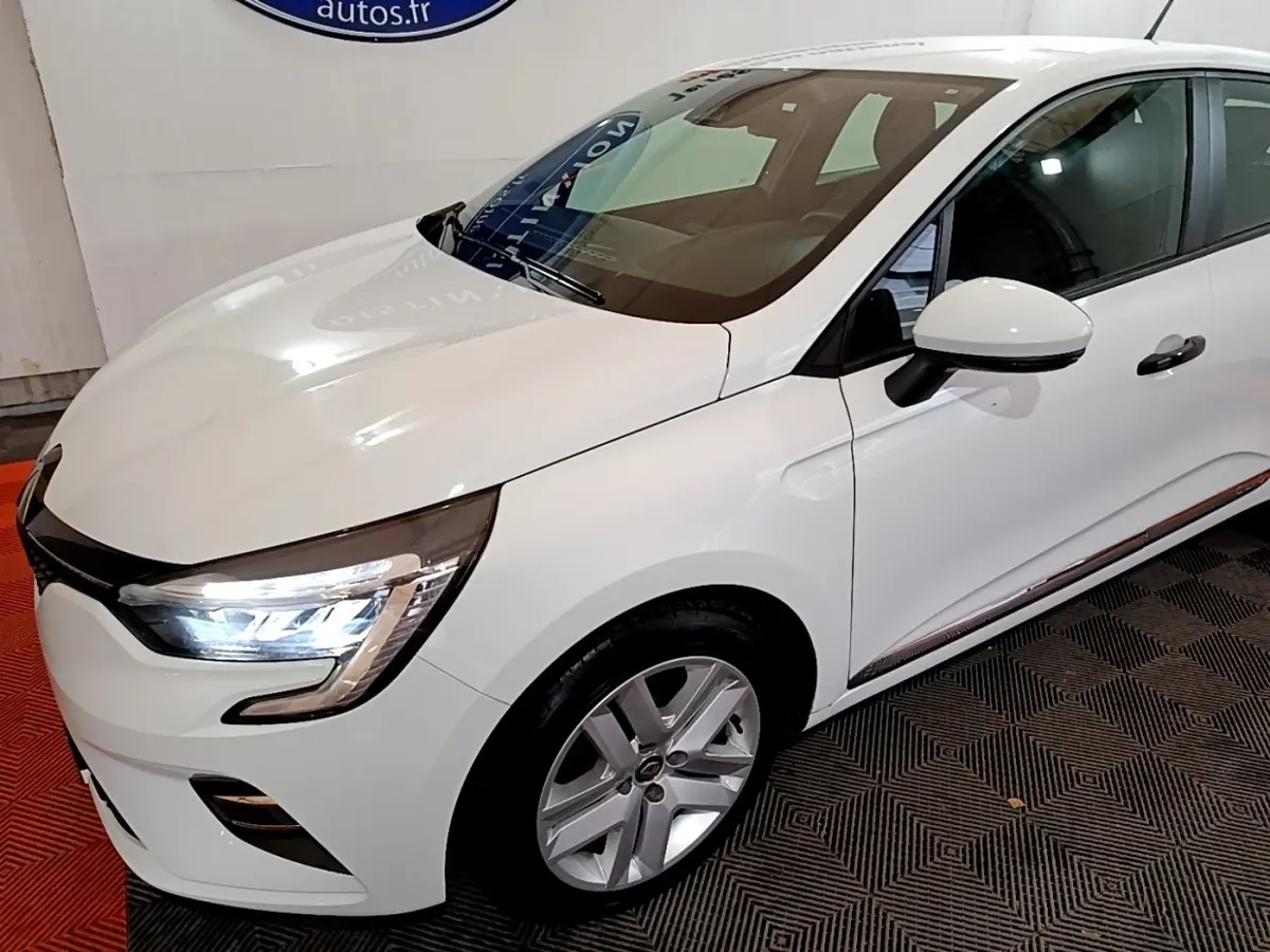 Vue 3/4 avant droit d'une Renault Clio Business blanche avec phares LED allumés et jantes alliage visibles.