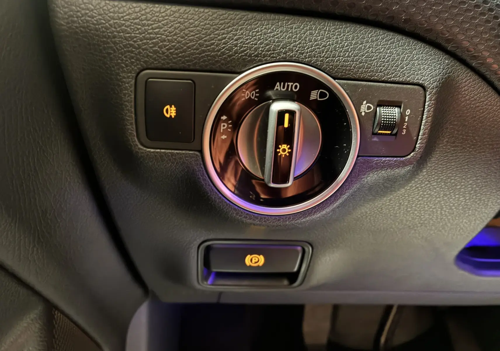 Gros plan sur le bouton de commande des phares et frein à main électronique dans l'habitacle noir d'une Mercedes Classe A 200D blanche de 2016.