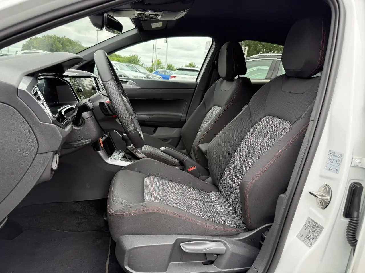 Intérieur avant droit d'une Volkswagen Polo blanche 2024, sièges tissu noir à motif écossais avec surpiqûres rouges.