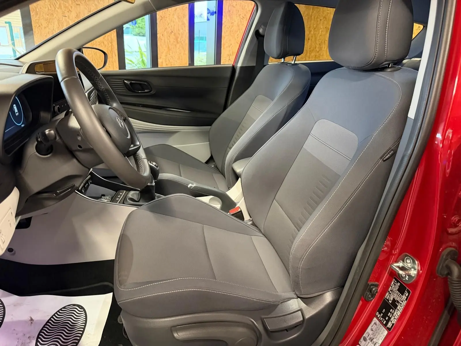 Vue intérieure côté conducteur du Hyundai Bayon rouge 2025, sièges tissu gris et volant cuir multifonctions.