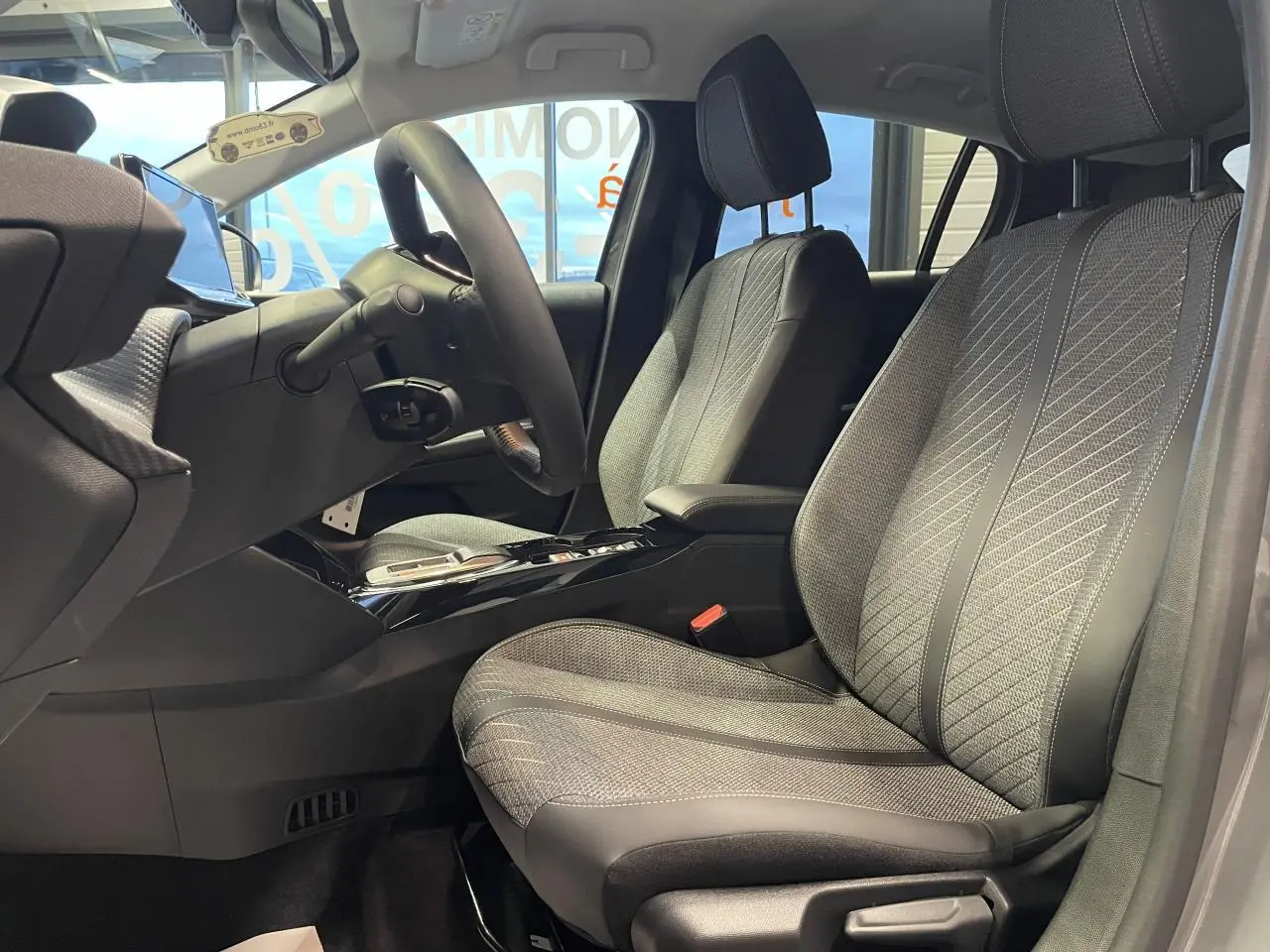 Vue intérieure côté gauche des sièges avant gris clair et noir de la Peugeot 208 1.2i Hybrid 2025 avec accoudoir central.