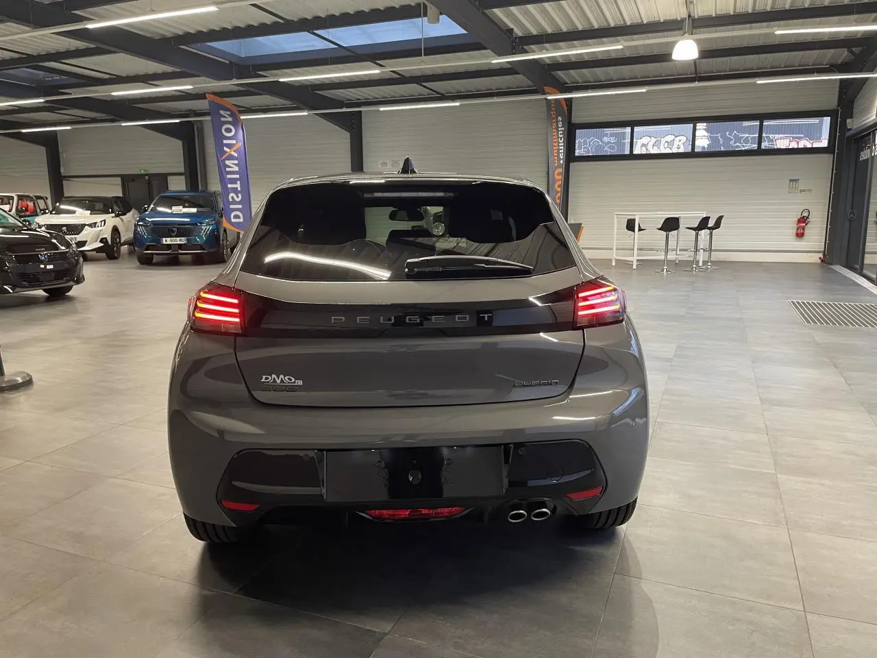 Vue arrière d'une Peugeot 208 hybride gris selenium avec feux arrière à LED allumés en intérieur showroom.